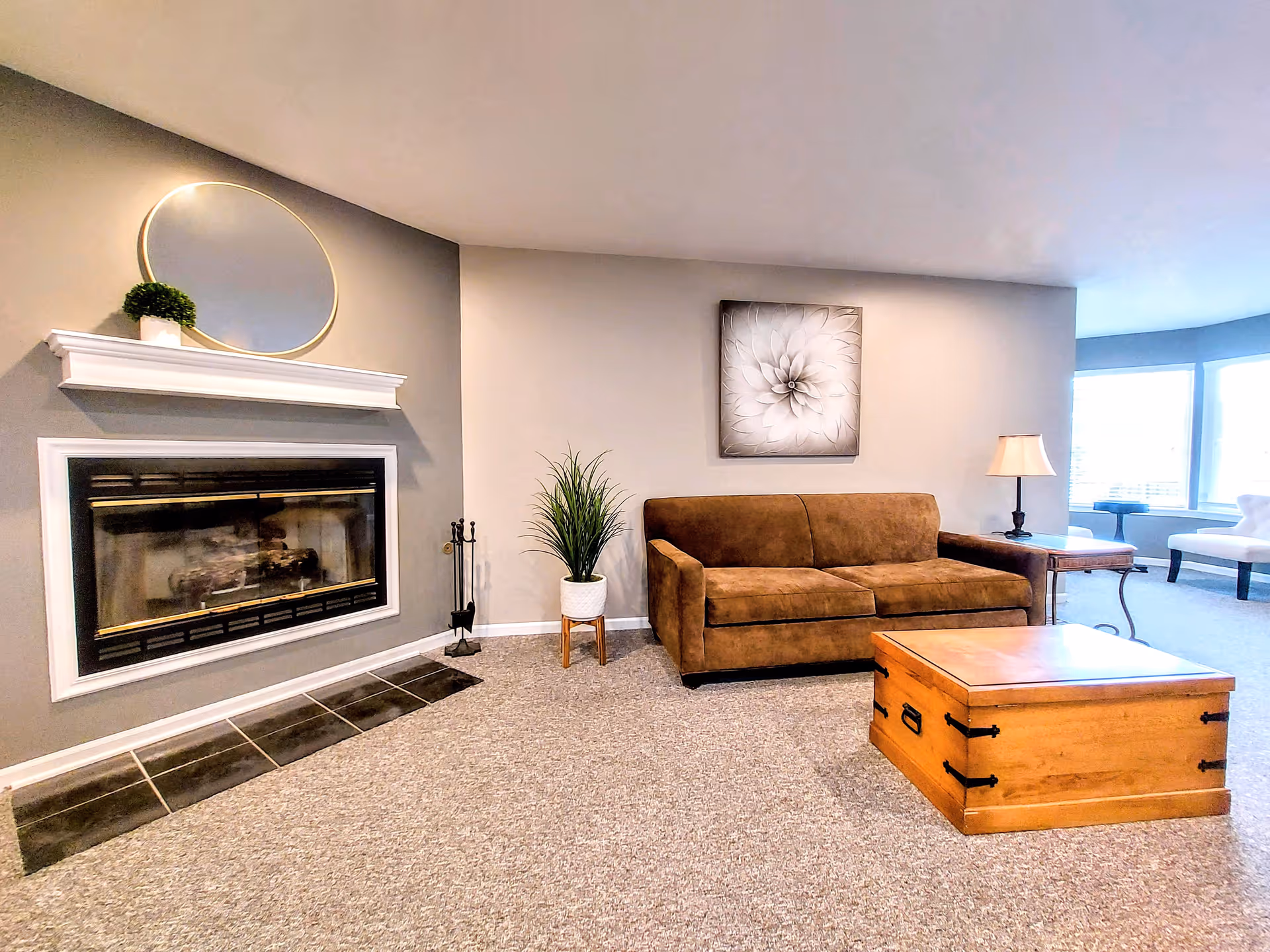 A cozy living room with a brown sofa, a wooden chest coffee table, a side table with a lamp, a potted plant, a fireplace with a white mantel and an oval mirror above it, and a large window letting in natural light.
