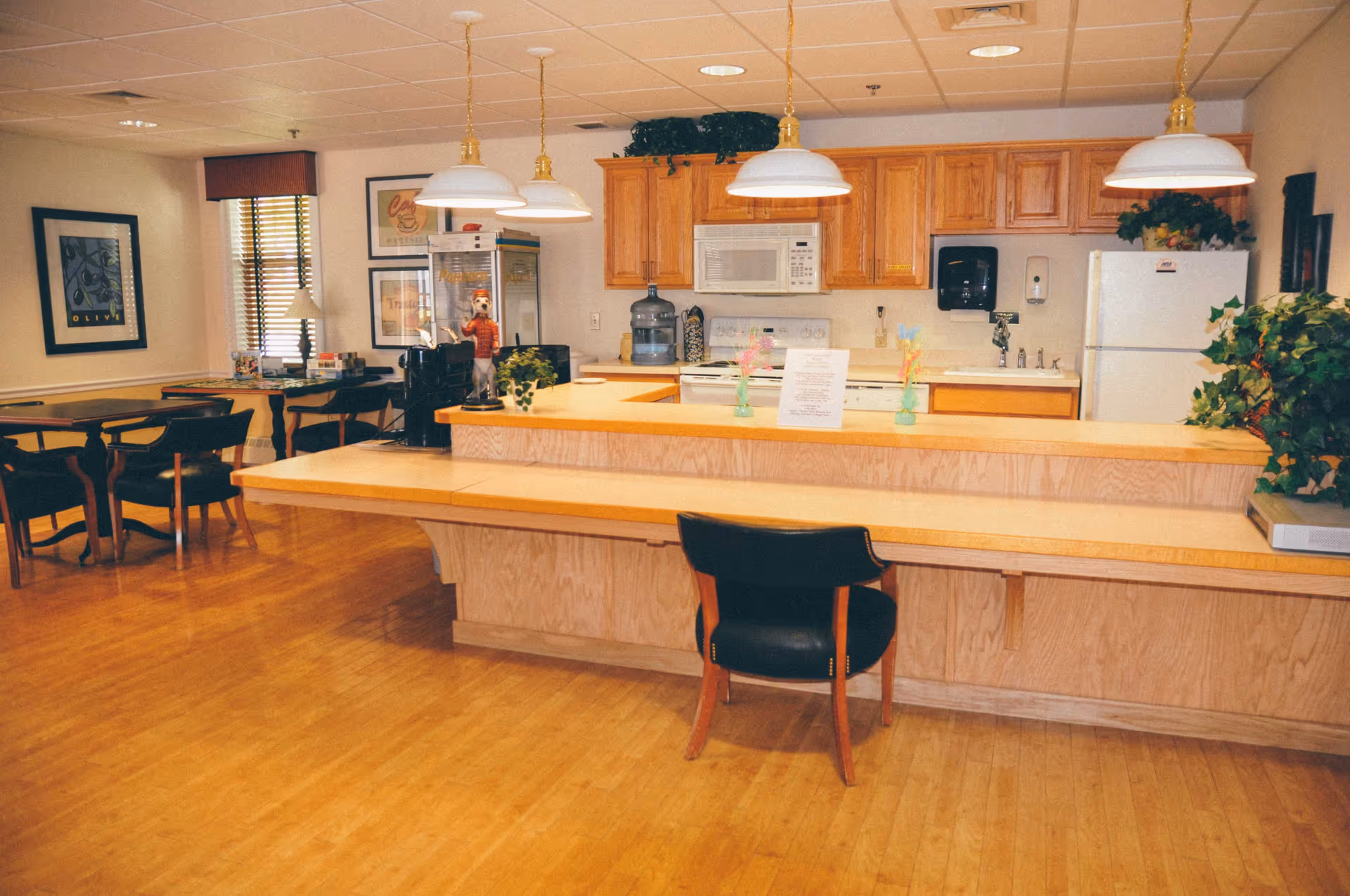 Communal dining and kitchenette area with a long wooden serving counter, chairs, tables, and hanging pendant lights.