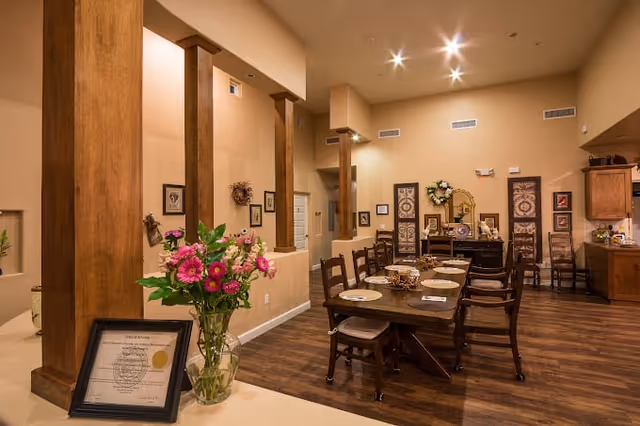 A warmly lit dining area with a long wooden table set with plates and napkins, surrounded by wooden chairs. The room features wooden pillars, framed artwork on the walls, and a bouquet of pink and purple flowers in a glass vase on a counter in the foreground. The flooring is wood, and there are decorative items on a sideboard against the far wall.