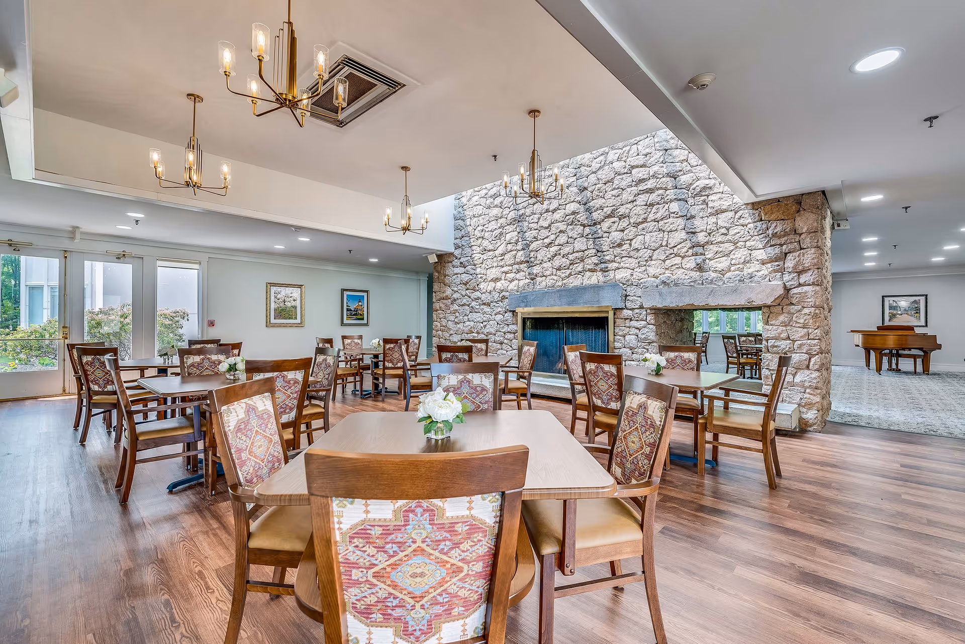 Spacious dining room with wooden tables and patterned chairs, a large stone fireplace, chandeliers, and windows letting in natural light.
