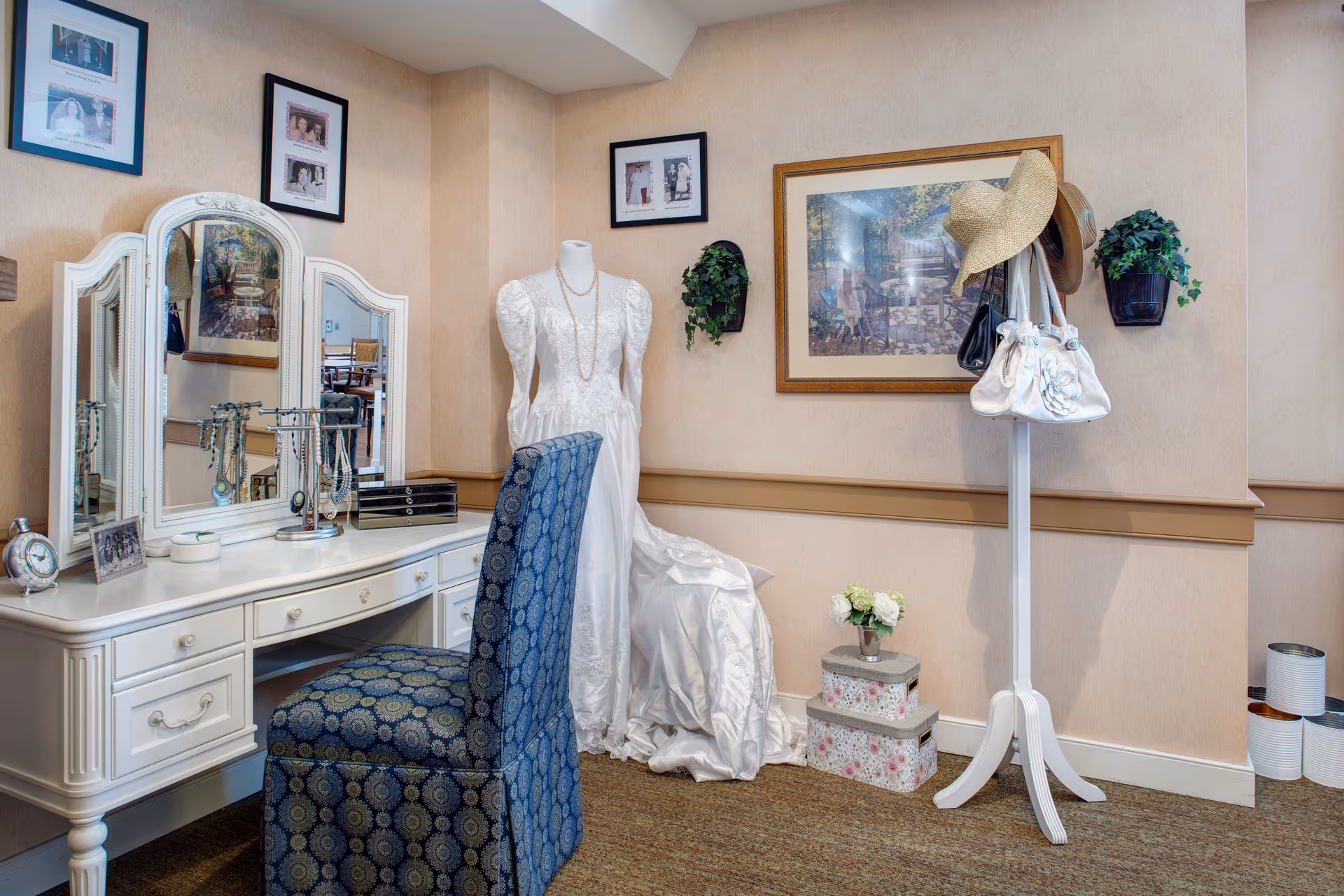 A cozy dressing area featuring a white vanity table with a three-panel mirror and a blue patterned chair. On the vanity are jewelry stands, a clock, and framed photos. Next to the vanity is a mannequin dressed in a white wedding gown with a pearl necklace. A white coat rack holds hats and handbags. The walls are decorated with framed pictures and small wall-mounted plants. Two floral storage boxes with a small flower arrangement sit on the floor.