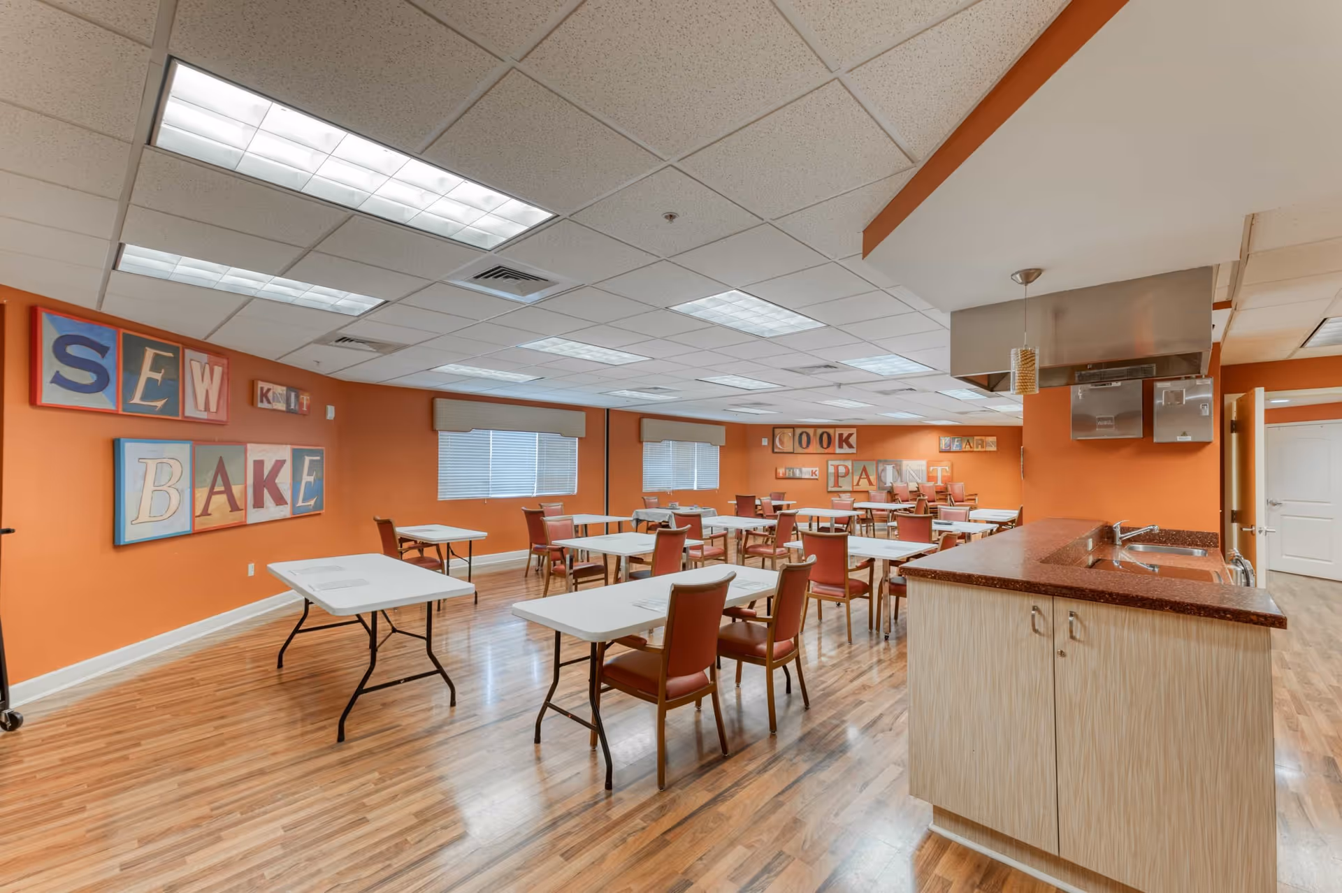 A spacious activity room with orange walls and wood flooring, featuring multiple tables and chairs arranged for group activities. The walls have colorful framed letters spelling out words like SEW, BAKE, COOK, PAINT, and LEARN. There is a kitchen counter with a sink and overhead ventilation hood on the right side of the room.