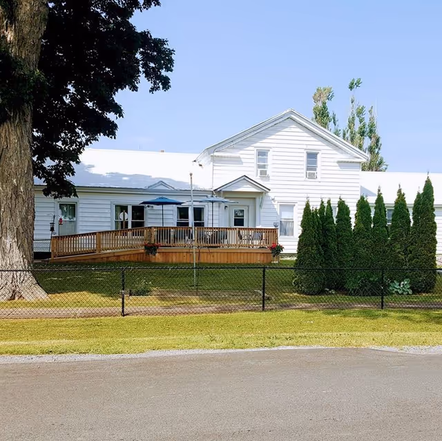 Exterior view of a white two-story building with a wooden deck and railing. The deck has a blue umbrella and some potted plants. There is a large tree on the left side and a row of tall evergreen shrubs on the right. A black chain-link fence runs along the front of the property, with a grassy area between the fence and the building. The sky is clear and blue.