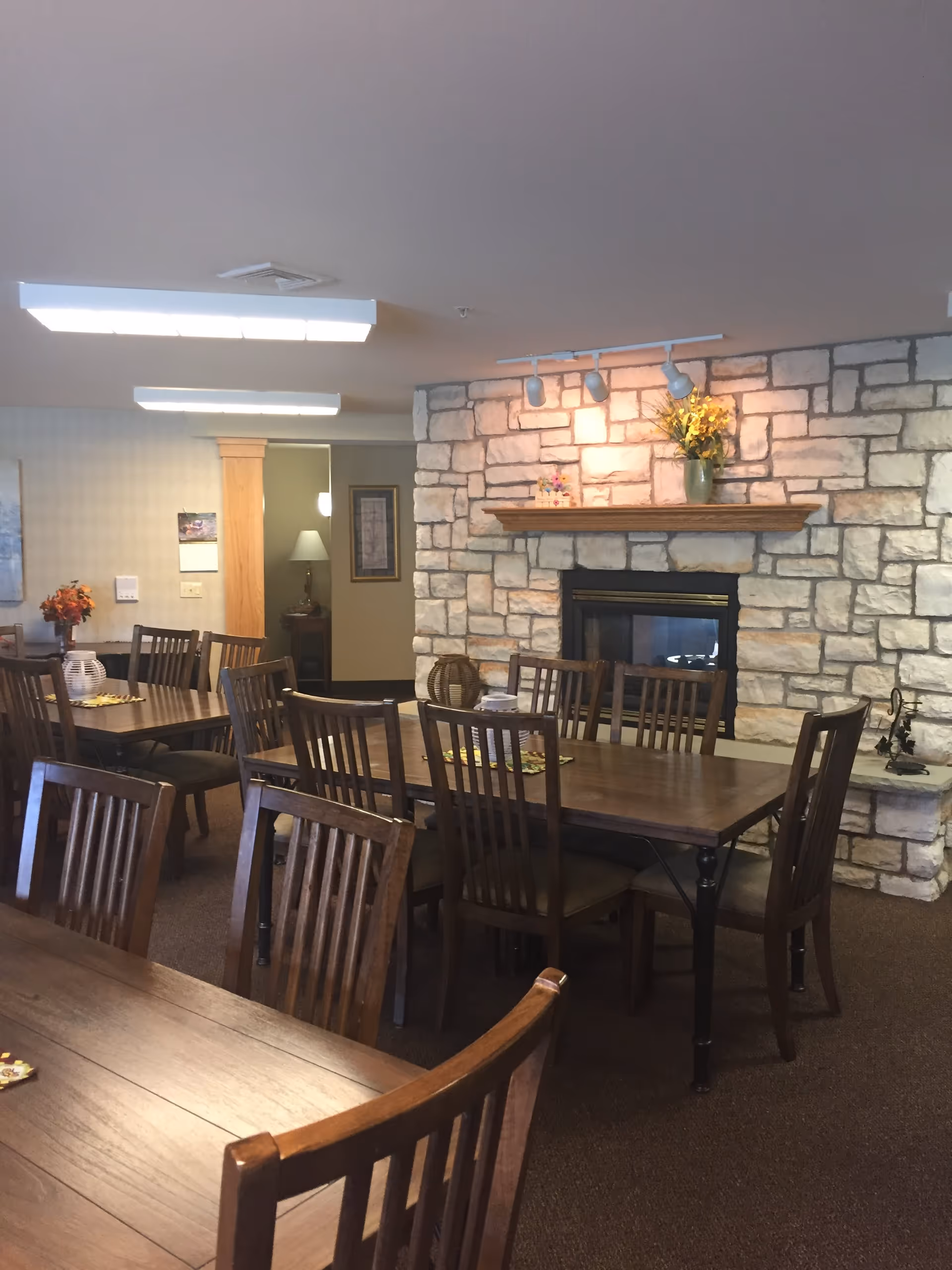 Interior view of a dining room with multiple wooden tables and chairs arranged neatly. A stone fireplace with a wooden mantle is decorated with a vase of yellow flowers and a small decorative item. The room is softly lit with ceiling lights and track lighting above the fireplace. There is a doorway leading to another room with a lamp and framed artwork visible.
