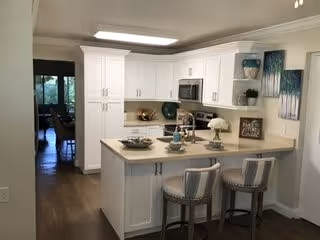 Bright and modern kitchen with white cabinets, a countertop island with two cushioned bar stools, and various decorative items including a lamp, bowls, and artwork on the walls. The kitchen opens into a dining area visible in the background.