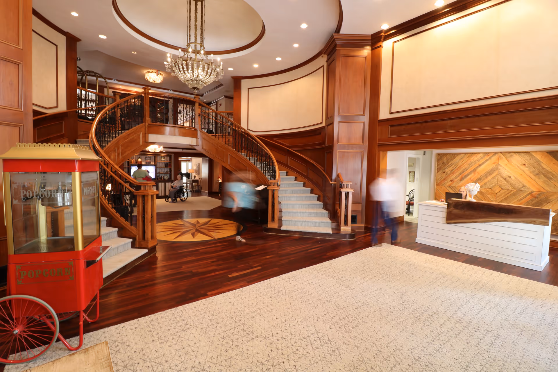 Grand interior lobby with a curved wooden staircase, chandelier, reception desk, and a red popcorn cart.