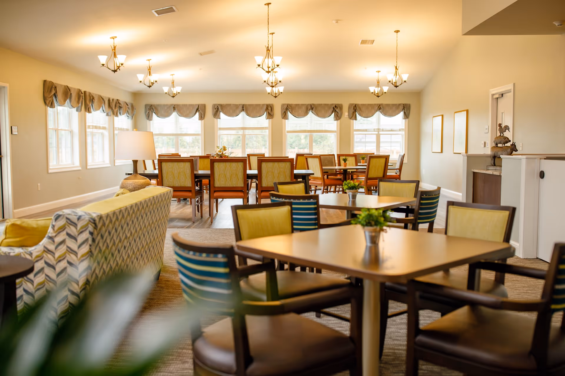 A bright and spacious dining room in an assisted living facility with multiple tables and chairs arranged neatly. The room has large windows with valances, several chandeliers hanging from the ceiling, and a cozy seating area with patterned sofas and a lamp.