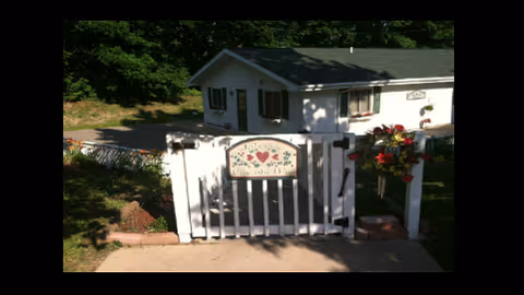 White wooden gate with a decorative sign featuring hearts and flowers, leading to a white house with black shutters surrounded by greenery and flowers.