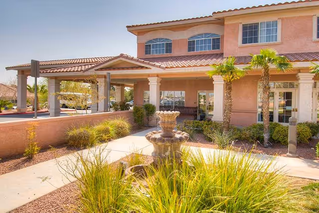 Exterior view of a two-story senior living facility with a covered entrance, palm trees, shrubs, and a decorative stone fountain in the foreground under a clear sky.