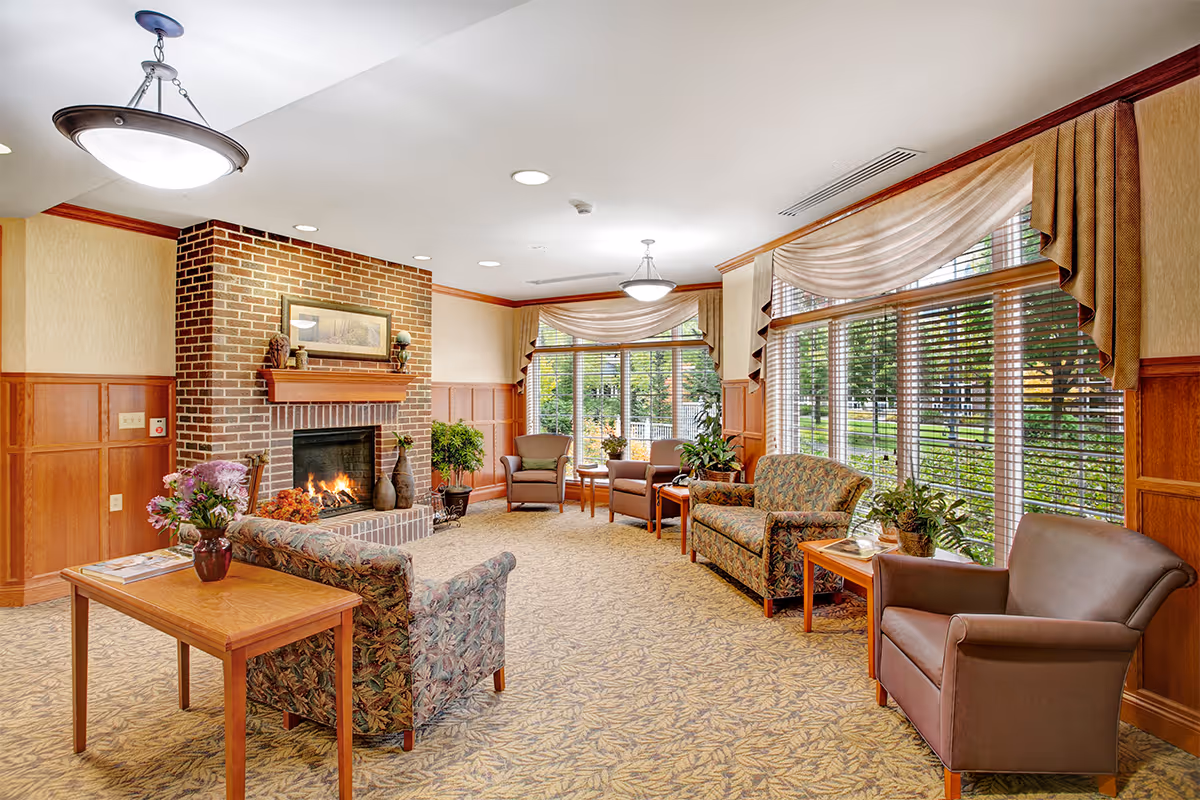 Well-lit senior living lounge with upholstered chairs and sofas arranged around a brick fireplace and large windows overlooking greenery.