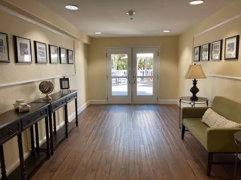 A well-lit interior hallway with wooden flooring leading to glass double doors that open to an outdoor patio area. The hallway features a green upholstered bench with a pillow on the right, a small round side table with a lamp, and a long console table on the left adorned with decorative items and framed black-and-white photos on the walls.