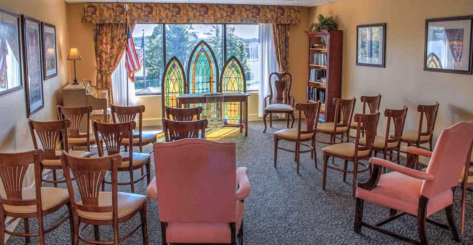 A small meeting or chapel room with several wooden chairs arranged in rows facing a table with three stained glass panels in front of a large window. The room has carpeted floors, framed pictures on the walls, a bookshelf, and an American flag near the window. There are two upholstered armchairs in the foreground.