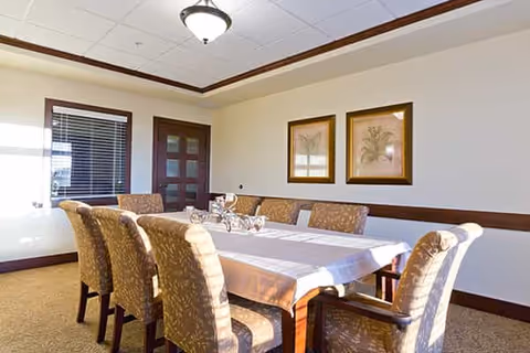 Dining room with a long table covered by a white tablecloth surrounded by upholstered chairs, framed artwork on the wall and a window with blinds.