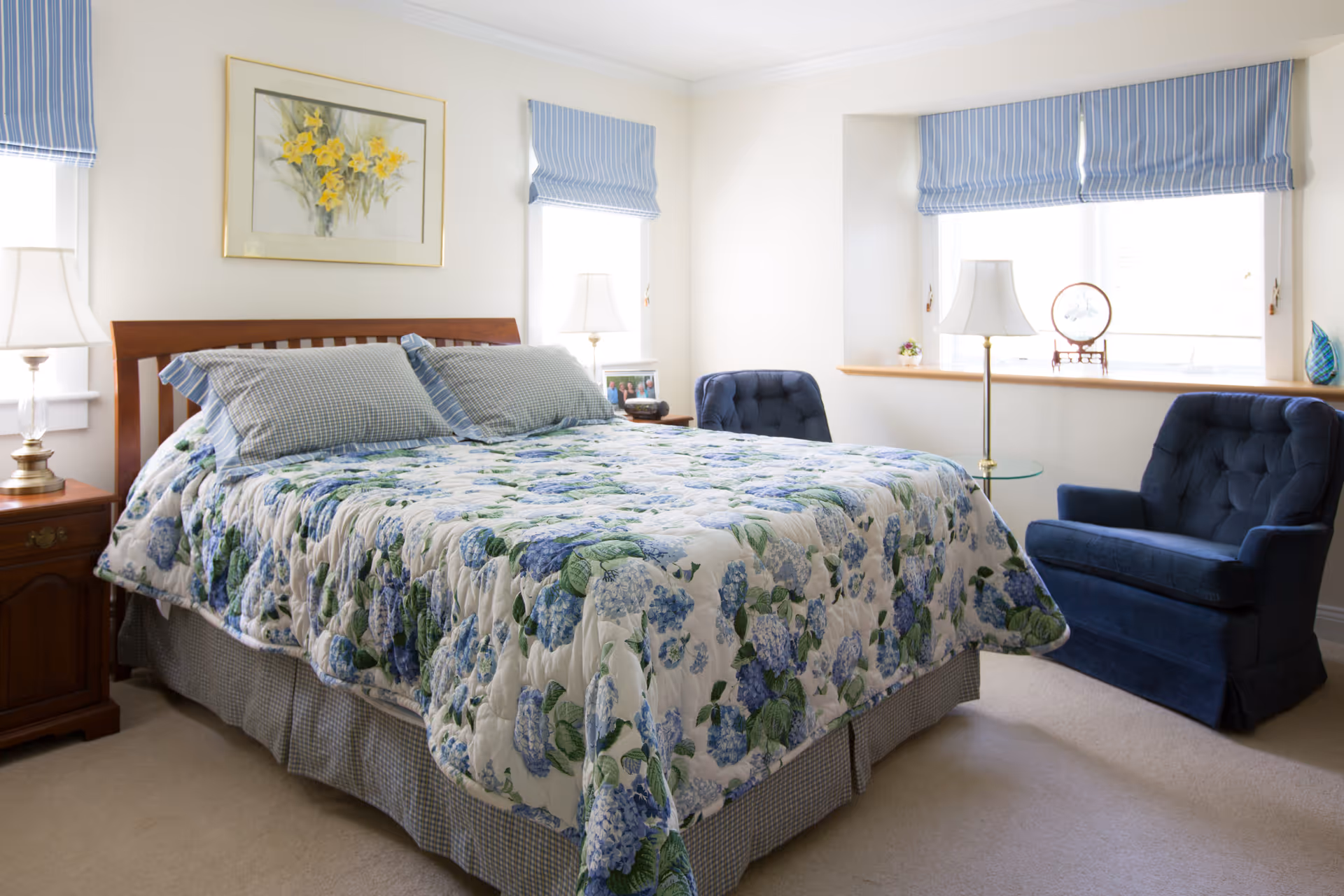 A bright bedroom with a wooden bed frame, a floral quilt with blue and green flowers, two blue checkered pillows, two bedside tables with lamps, two blue upholstered armchairs, and windows with blue striped Roman shades. A framed painting of yellow flowers hangs above the bed.