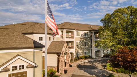 Exterior front of a multi-story senior living building with an American flag, driveway, and landscaping under a partly cloudy sky.