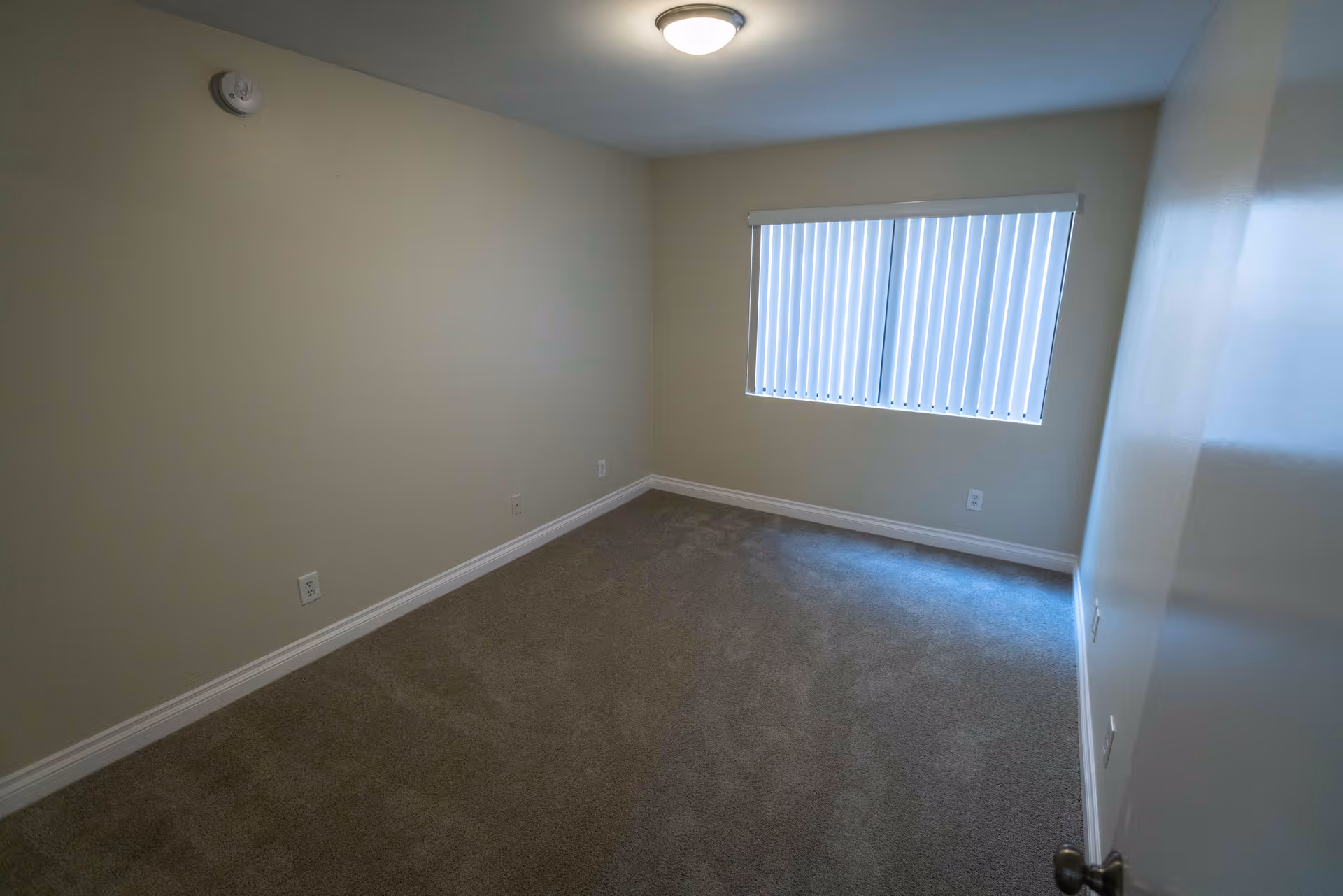 Empty room with beige walls, carpeted floor, a window with vertical blinds, a ceiling light fixture, and a door partially open with a round doorknob.