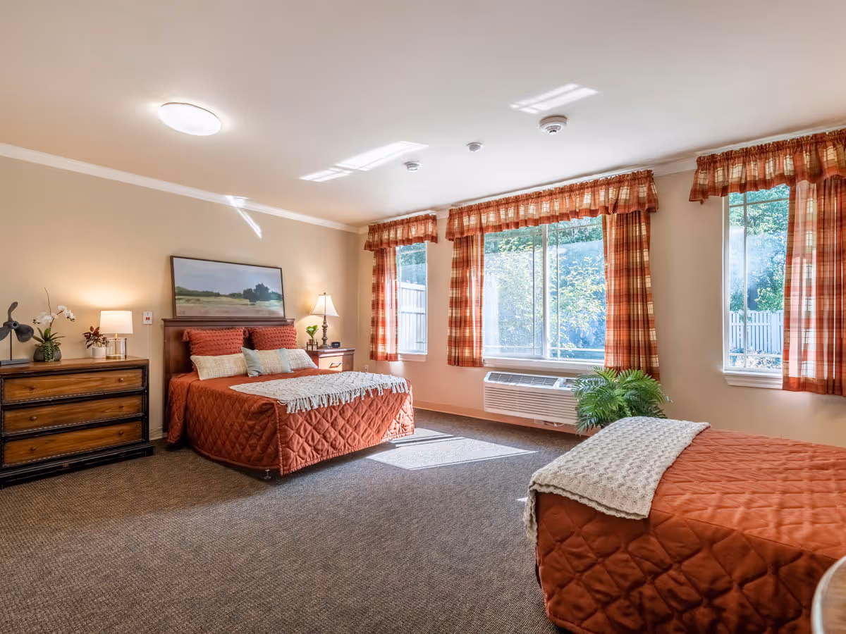 A bright bedroom with two beds covered in rust-colored quilted bedspreads and white knitted throws. The room has beige walls, a carpeted floor, and three large windows with plaid curtains letting in natural light. There is a wooden dresser with plants and lamps on it, and a framed landscape painting above the headboard of one bed.