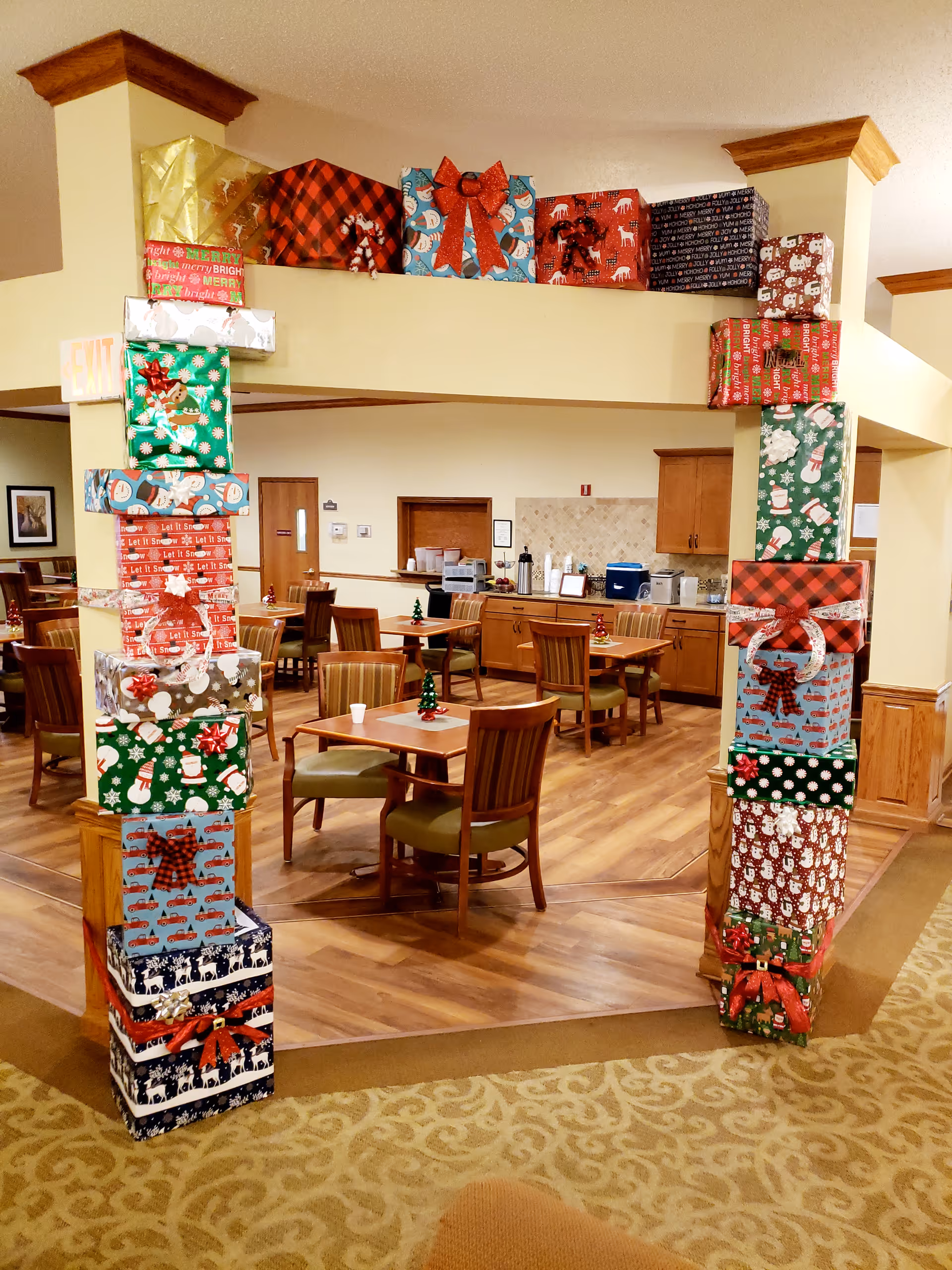 Interior view of a dining area decorated for the holidays with a large archway made of wrapped Christmas presents. The room has wooden floors, tables with chairs, and a kitchen area in the background with cabinets and appliances.