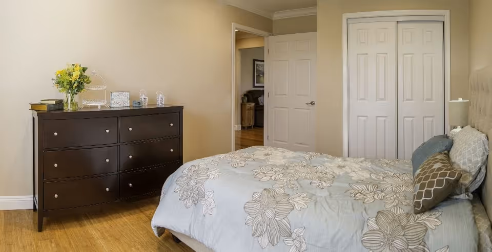 A bedroom with a bed covered in a floral patterned comforter and several pillows. To the left is a dark wooden dresser with books, a vase of yellow flowers, and decorative items on top. The room has light-colored walls and wooden flooring. There is a white door slightly open leading to another room and a closed closet with double doors.