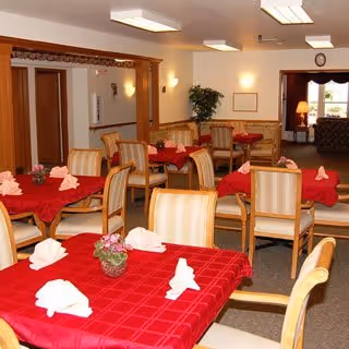 Dining room with multiple tables covered in red tablecloths, each set with folded white napkins and small flower centerpieces. Wooden chairs with cushioned seats surround the tables. The room has warm lighting, a plant in the corner, and an open doorway leading to another seating area with a lamp and sofa.