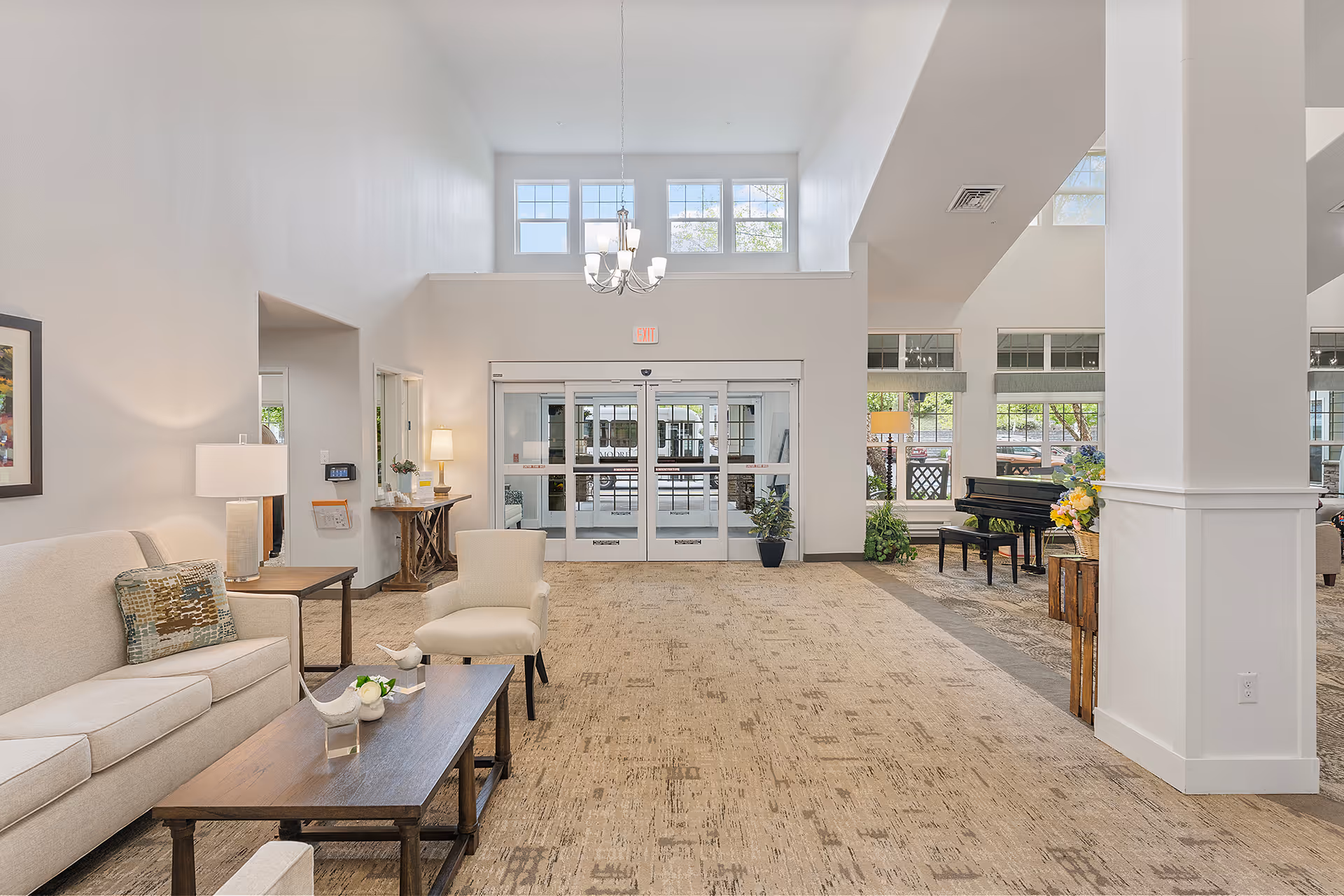 Bright, spacious retirement community lobby with seating, a coffee table, chandelier, and glass entrance doors.