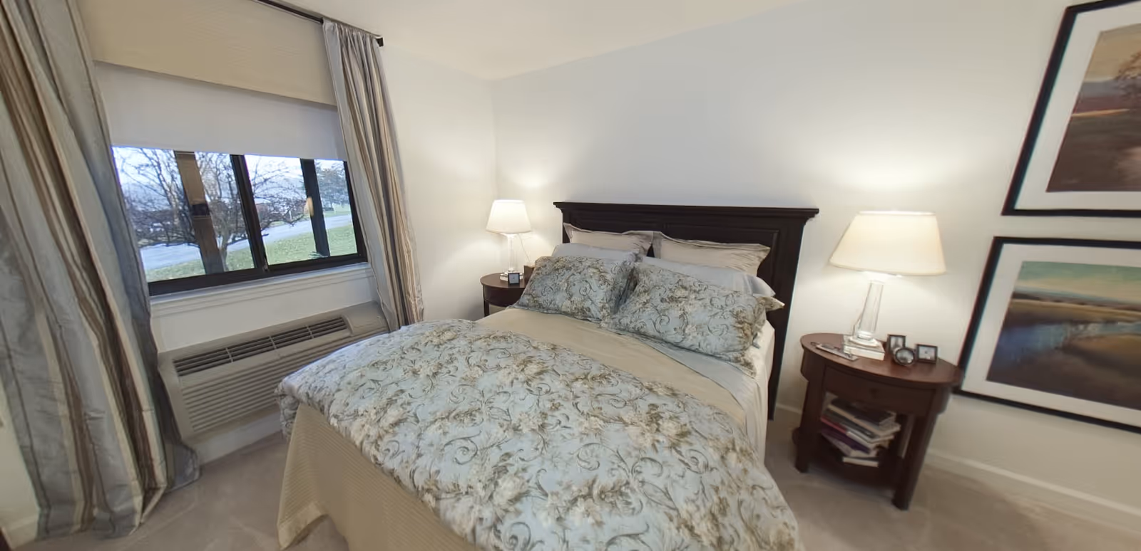 A neatly made bed with floral patterned bedding and multiple pillows in a bedroom. There are two wooden nightstands on either side of the bed, each with a lamp. The nightstand on the right also has some small picture frames and books on the lower shelf. A window with curtains and a wall-mounted air conditioning unit is visible to the left. Two framed landscape paintings hang on the wall to the right.