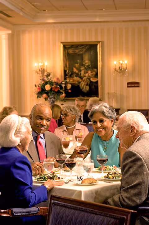 Four elderly people sitting around a dining table in a warmly lit room, enjoying a meal together with wine glasses and plates of food. The background shows other diners, a floral arrangement, wall sconces, and a framed painting on striped wallpaper.