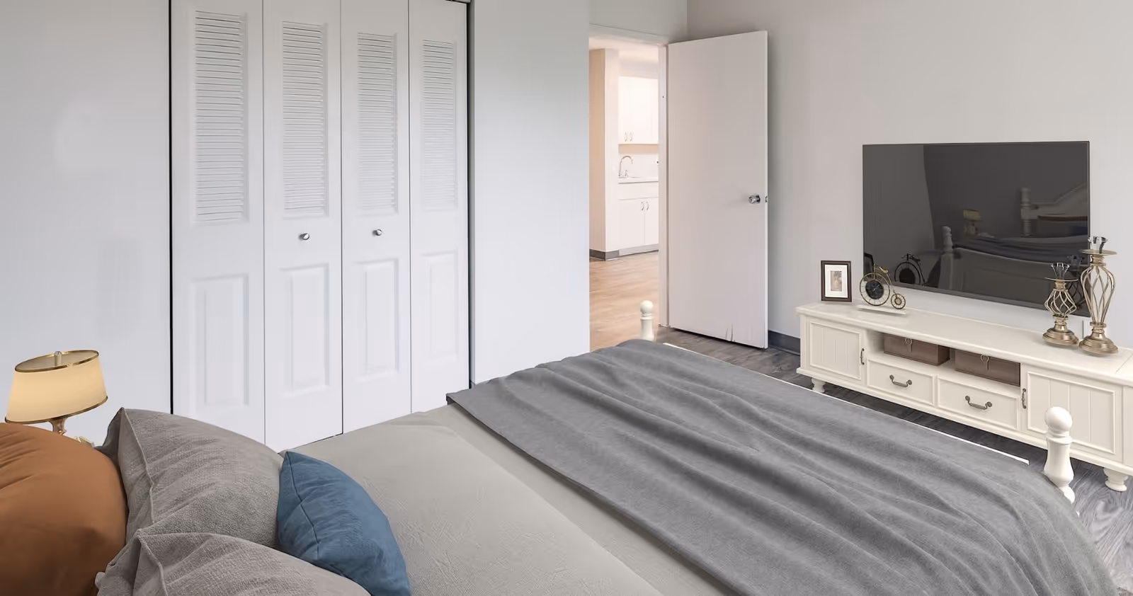 A bedroom with a bed covered in gray and beige bedding, a white TV stand with a flat-screen TV, decorative items, and a lamp. There is a closed white closet with louvered doors and an open door leading to a kitchen area.