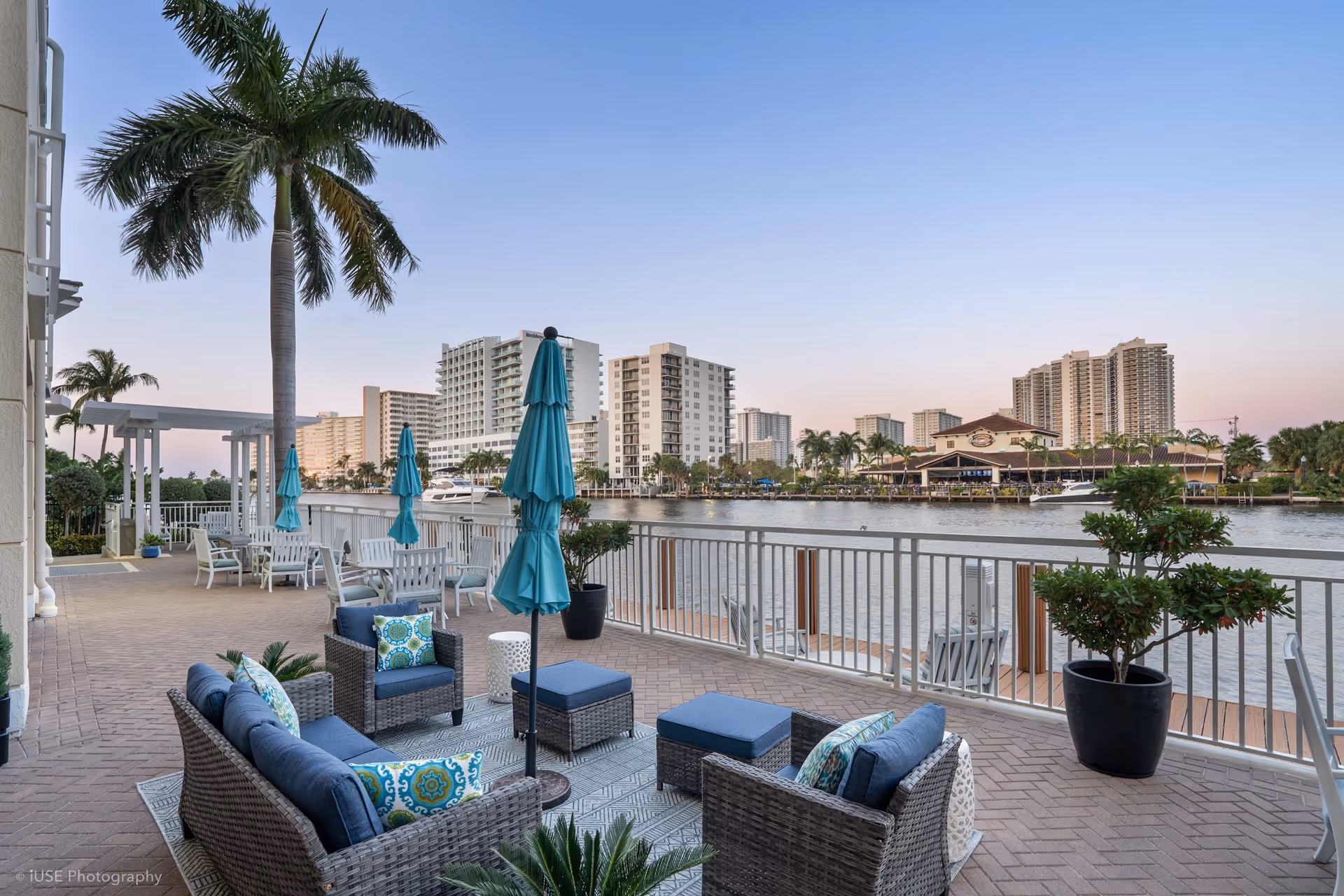Outdoor patio area at The Meridian at Waterways featuring wicker seating with blue cushions and patterned pillows, several closed turquoise umbrellas, palm trees, and a waterfront view with buildings across the water under a clear sky at dusk.