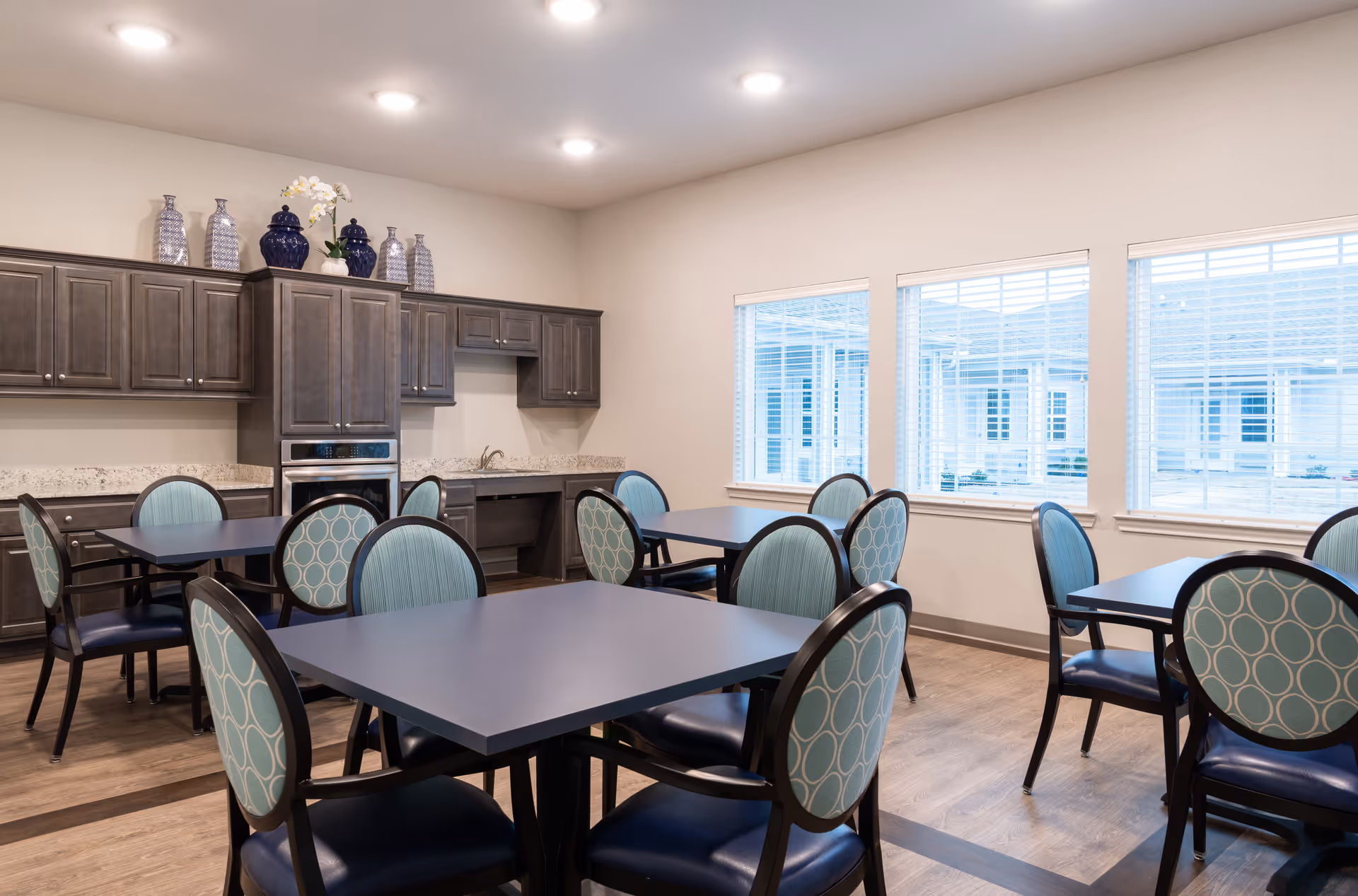 A bright dining room with several square tables and chairs featuring blue patterned upholstery. The room has large windows with white blinds allowing natural light to enter. Along one wall, there are dark wooden cabinets with decorative vases on top and a built-in oven beneath the countertop.