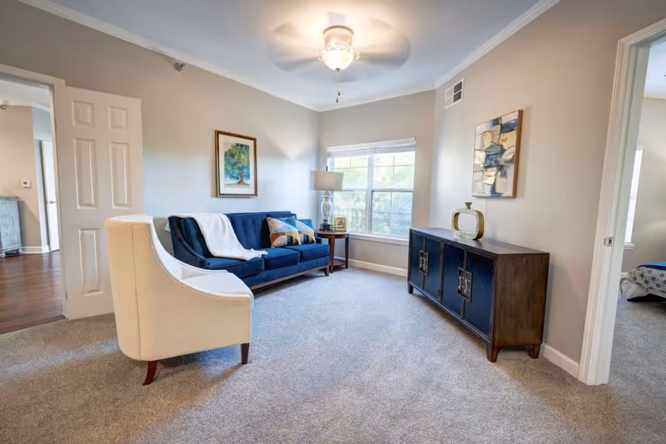 A bright living room with a blue sofa adorned with pillows and a white throw blanket, a beige armchair, a wooden side table with a lamp and picture frame, a large window letting in natural light, and a dark wooden cabinet with decorative items on top. The room has beige walls, carpeted floor, and two open doorways leading to other rooms.