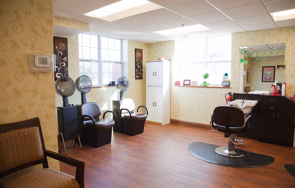 Bright hair salon room with styling chairs, hooded dryers, a shampoo sink and large windows in a senior living facility.