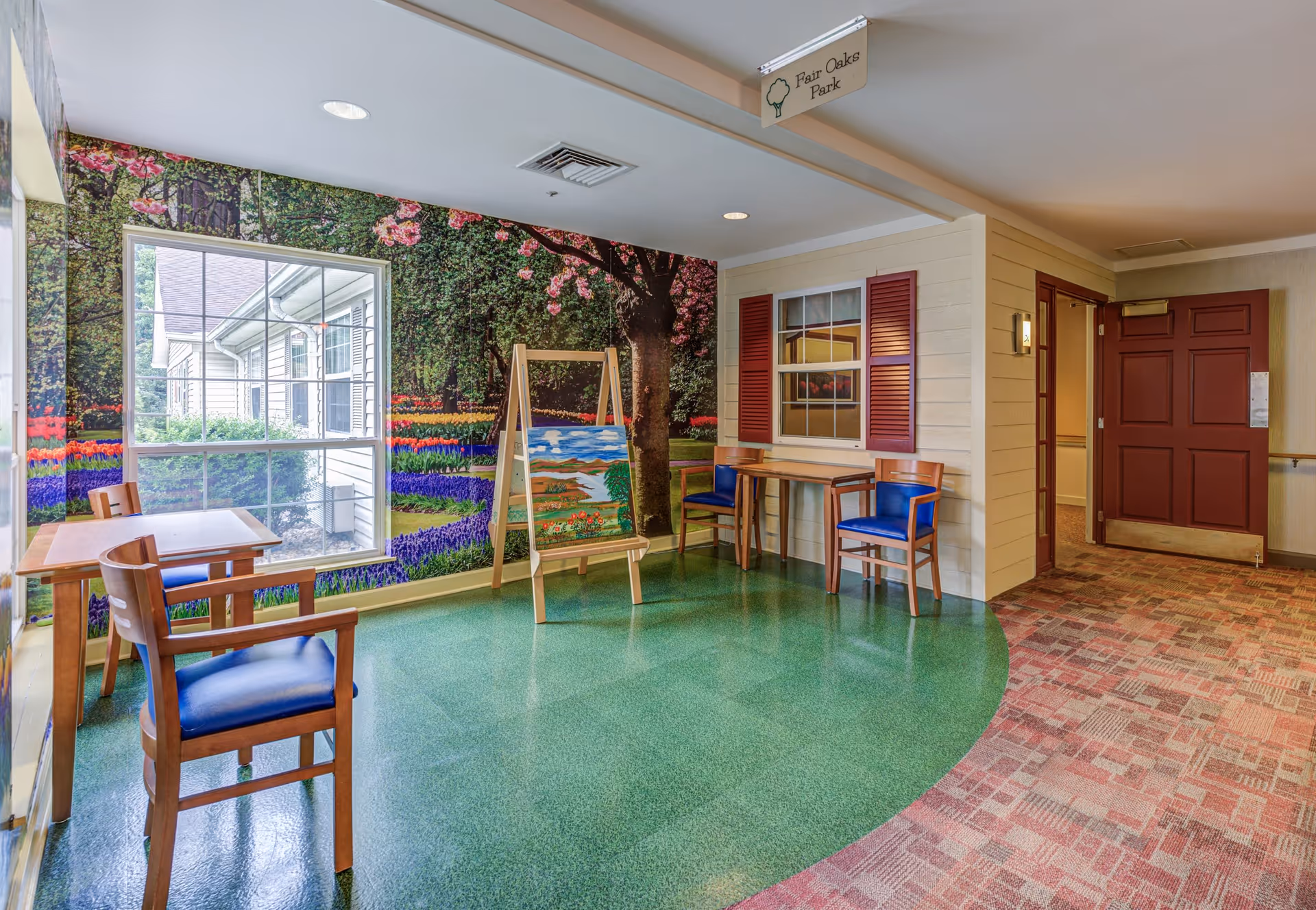 A bright indoor seating area with two wooden tables and four chairs with blue cushions. The walls feature a large mural of a colorful garden with flowers and trees, and a window showing the outside of the building. An easel with a painting is placed near the mural. The floor is a combination of green linoleum and patterned carpet. A sign above reads 'Fair Oaks Park'.