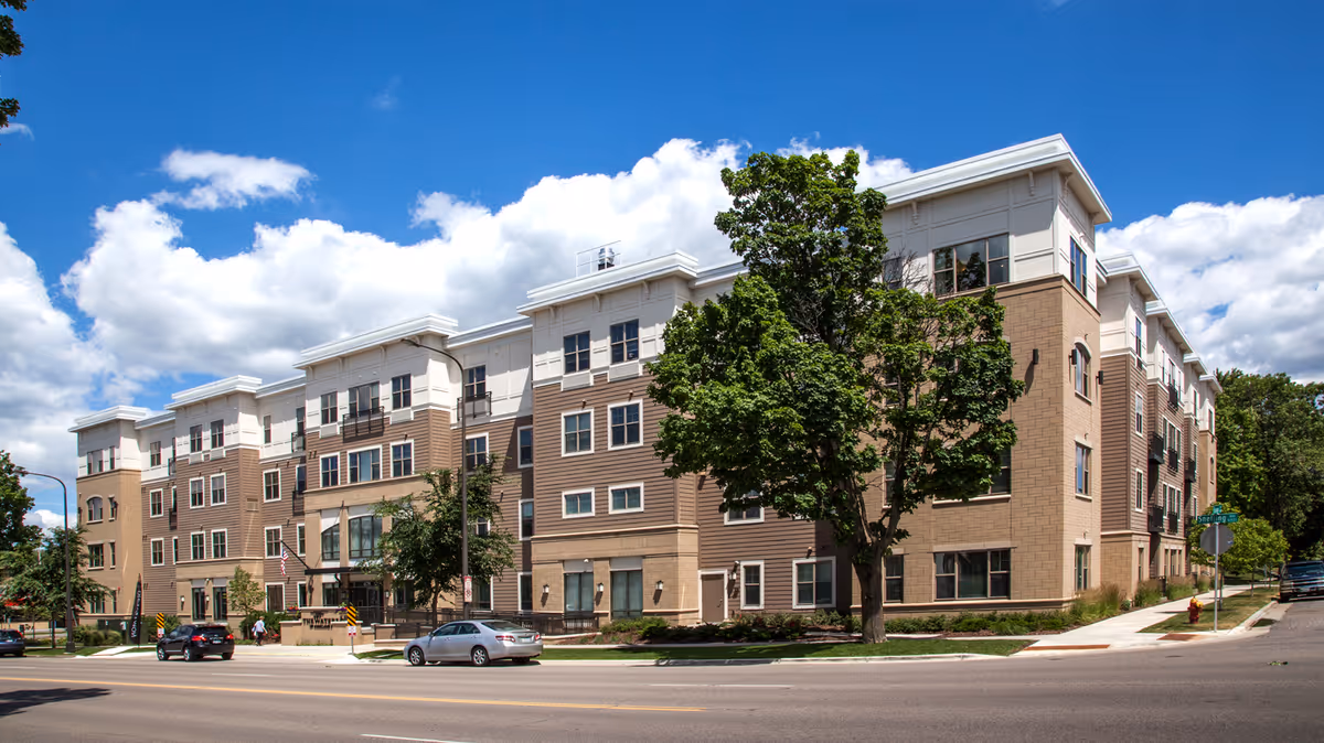 Exterior view of a multi-story senior living facility building with beige and brown siding, multiple windows, and a tree in front. Cars are parked along the street, and the sky is blue with scattered clouds.