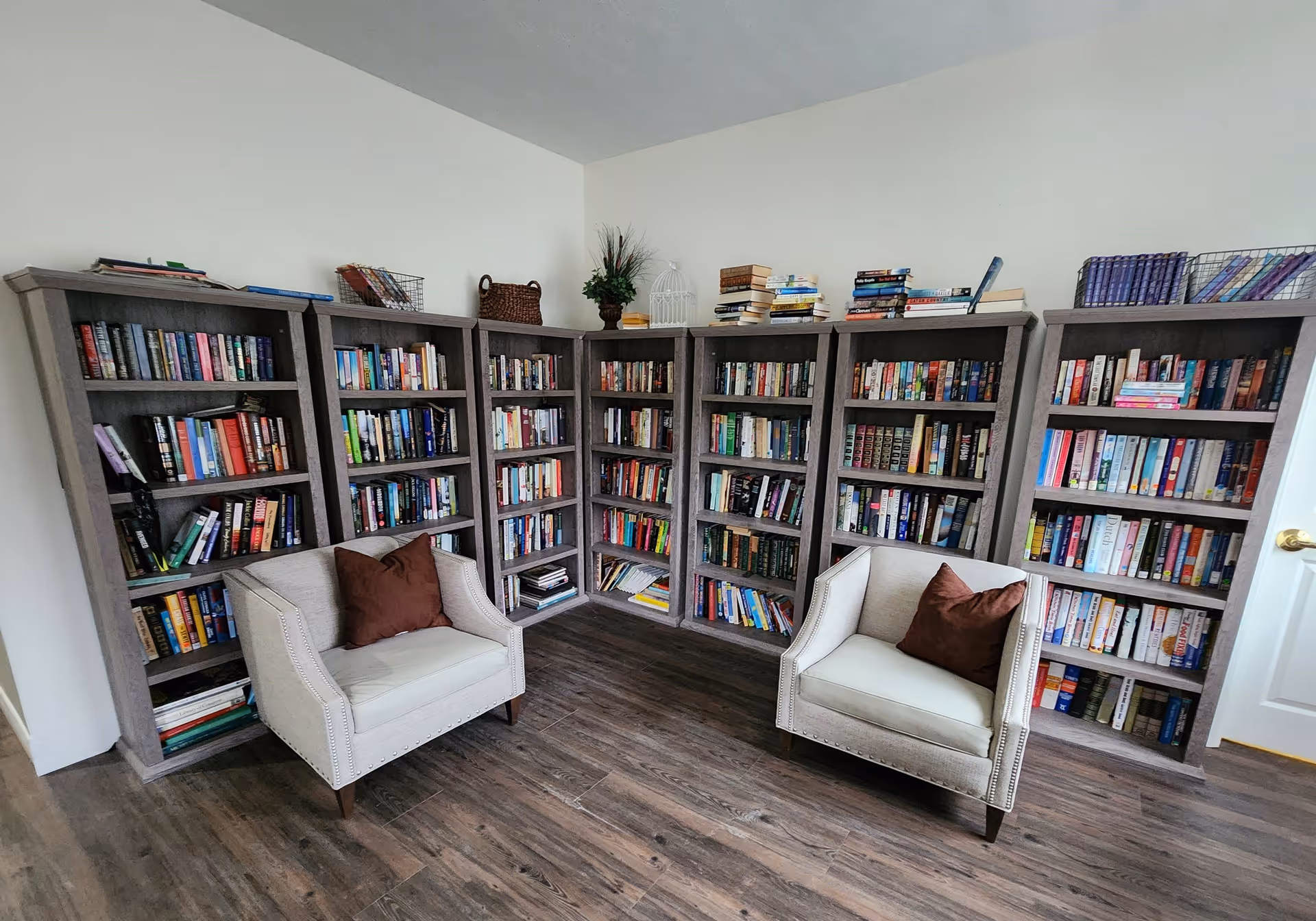 A cozy reading nook in a senior living facility featuring six tall bookshelves filled with books arranged along two walls. Two beige armchairs with brown cushions are placed facing each other on a wooden floor. Decorative items like baskets and plants are placed on top of the bookshelves.