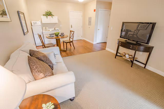 Bright living area with a beige sofa and throw pillows, a small round dining table with chairs, a flat-screen TV on a stand, and an entry door in the background.