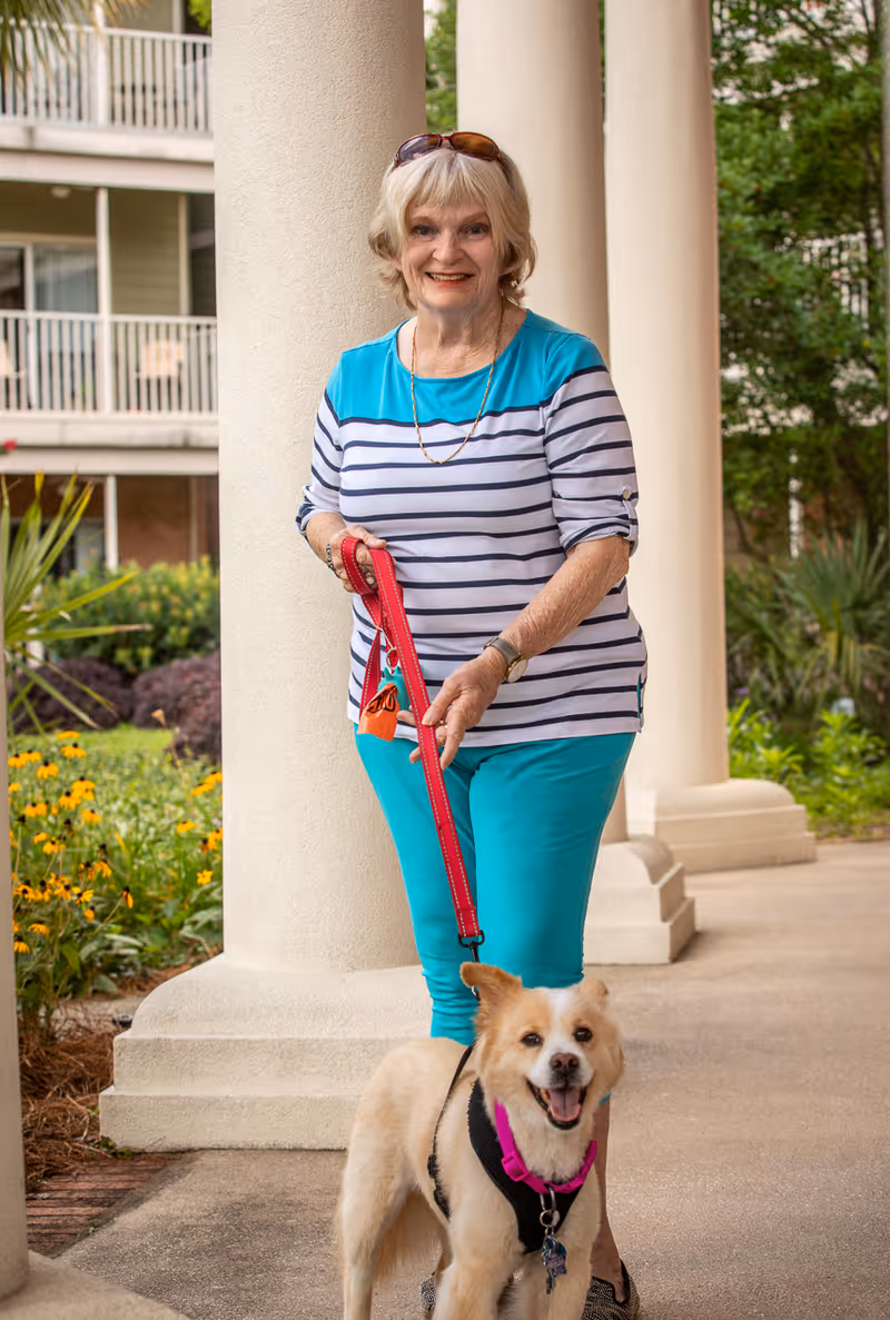 An elderly woman wearing a striped shirt and turquoise pants is standing outside near large white columns, holding a red leash attached to a happy medium-sized dog with light fur and a pink harness. There are green plants and flowers in the background along with a building with balconies.