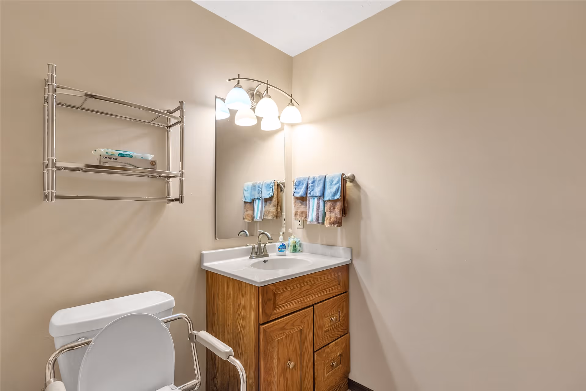 A bathroom with beige walls featuring a white toilet with metal safety rails, a wooden vanity with a white sink, a mirror above the sink, a light fixture with four bulbs, a towel rack with blue and brown towels, and a metal shelf holding a box of wipes.