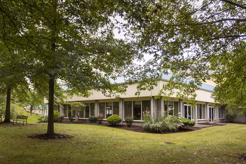 Exterior view of a single-story senior living facility building surrounded by green grass, trees, and shrubs. The building has large windows and a light-colored roof, with a bench visible on the left side under the trees.