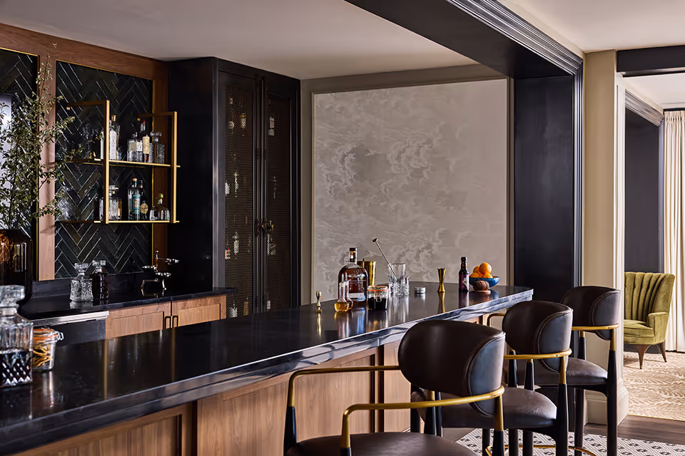 A modern bar area with a long black countertop and wooden cabinetry. There are several bottles and glassware on the counter and shelves, along with three black and gold bar stools. The background features a textured wall and a glimpse of a green armchair in an adjacent room.