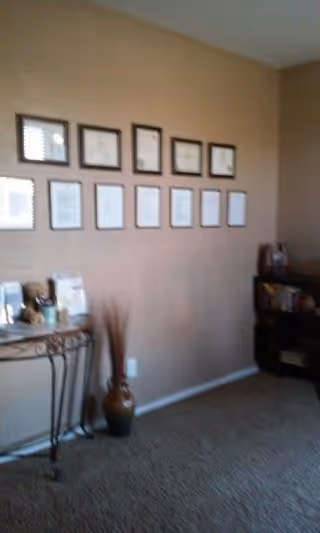 A beige wall with multiple framed certificates or documents arranged in two rows. Below the frames, there is a decorative table with various items on it and a tall vase with dried branches. To the right, there is a small bookshelf with books and other items. The floor is carpeted.