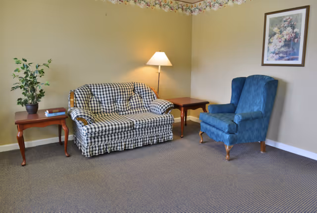 A small sitting area with a checkered loveseat, a blue upholstered armchair, two wooden side tables, a floor lamp, a potted plant, and a framed floral painting on the wall.
