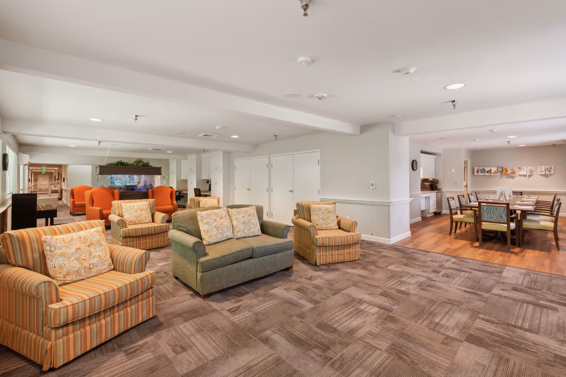 Spacious communal living area with upholstered armchairs and sofas on carpet, a dining table to the right, and an aquarium in the background.
