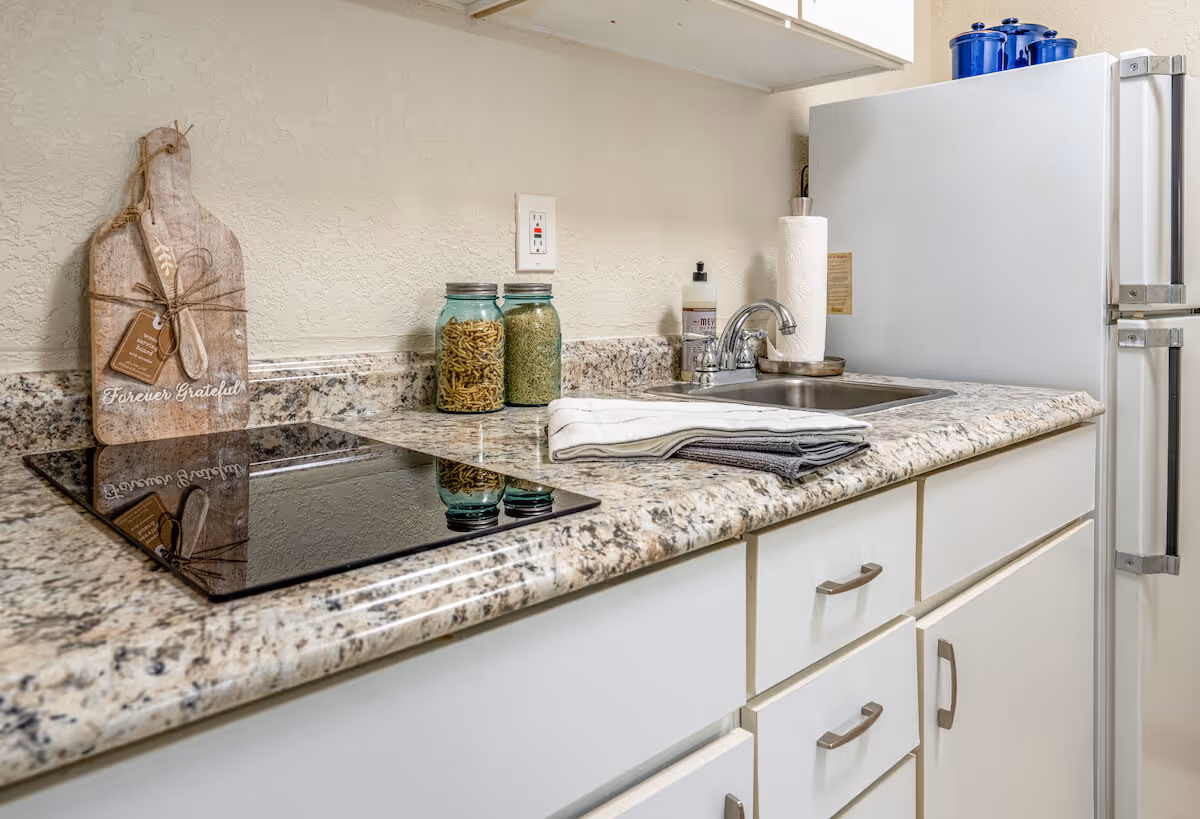 A clean kitchen countertop with a built-in electric stovetop, two glass jars filled with pasta and grains, a wooden cutting board with the words 'Forever Grateful', a sink with a faucet, paper towels, hand soap, and a white refrigerator in the background.