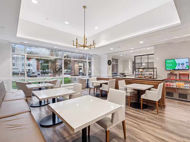 A bright and modern dining area with white tables and beige chairs, a long cushioned bench along one wall, large windows letting in natural light, and a counter with a display case and beverages. A chandelier hangs from the ceiling.
