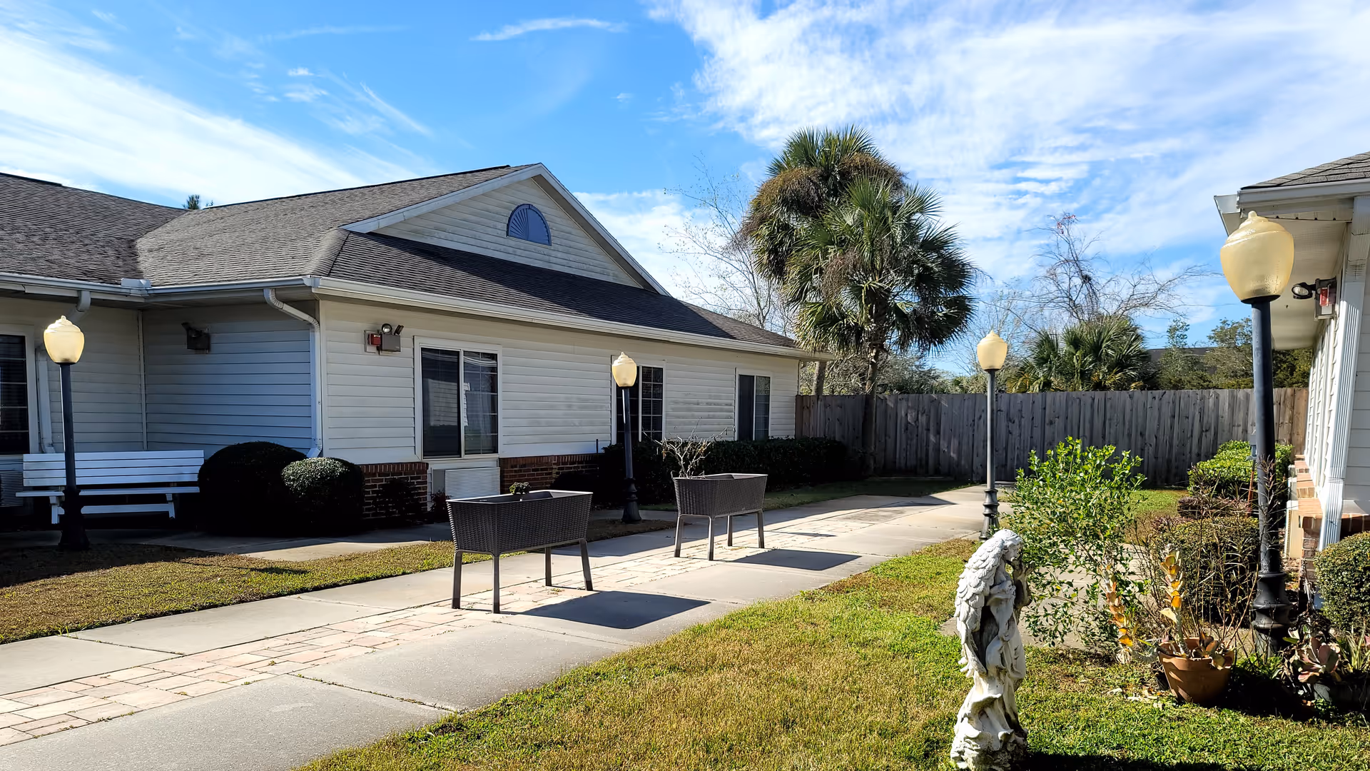 Sunny courtyard between single-story white residential buildings with benches, planters, lampposts and a small statue.