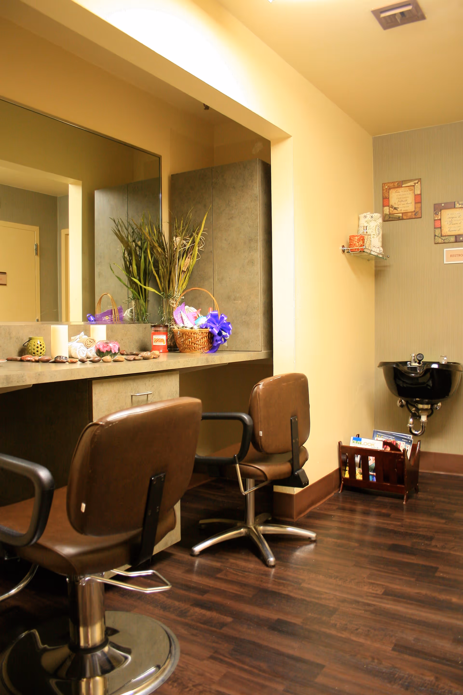 Interior view of a salon area with two brown salon chairs in front of a counter with a large mirror. The counter has decorative items including a basket with a purple ribbon, a candle, and some plants. There is a black wash basin mounted on the wall to the right and a small rack with magazines below it. The floor is dark wood and the walls are painted in neutral tones.