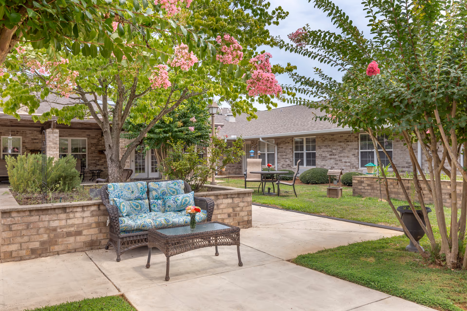 Outdoor patio area at Brookdale Eden featuring a wicker loveseat with blue patterned cushions and a matching wicker coffee table with a vase of flowers. The patio is surrounded by green grass, flowering trees with pink blossoms, and brick walls. In the background, there are additional tables and chairs near a brick building with windows and a roof.