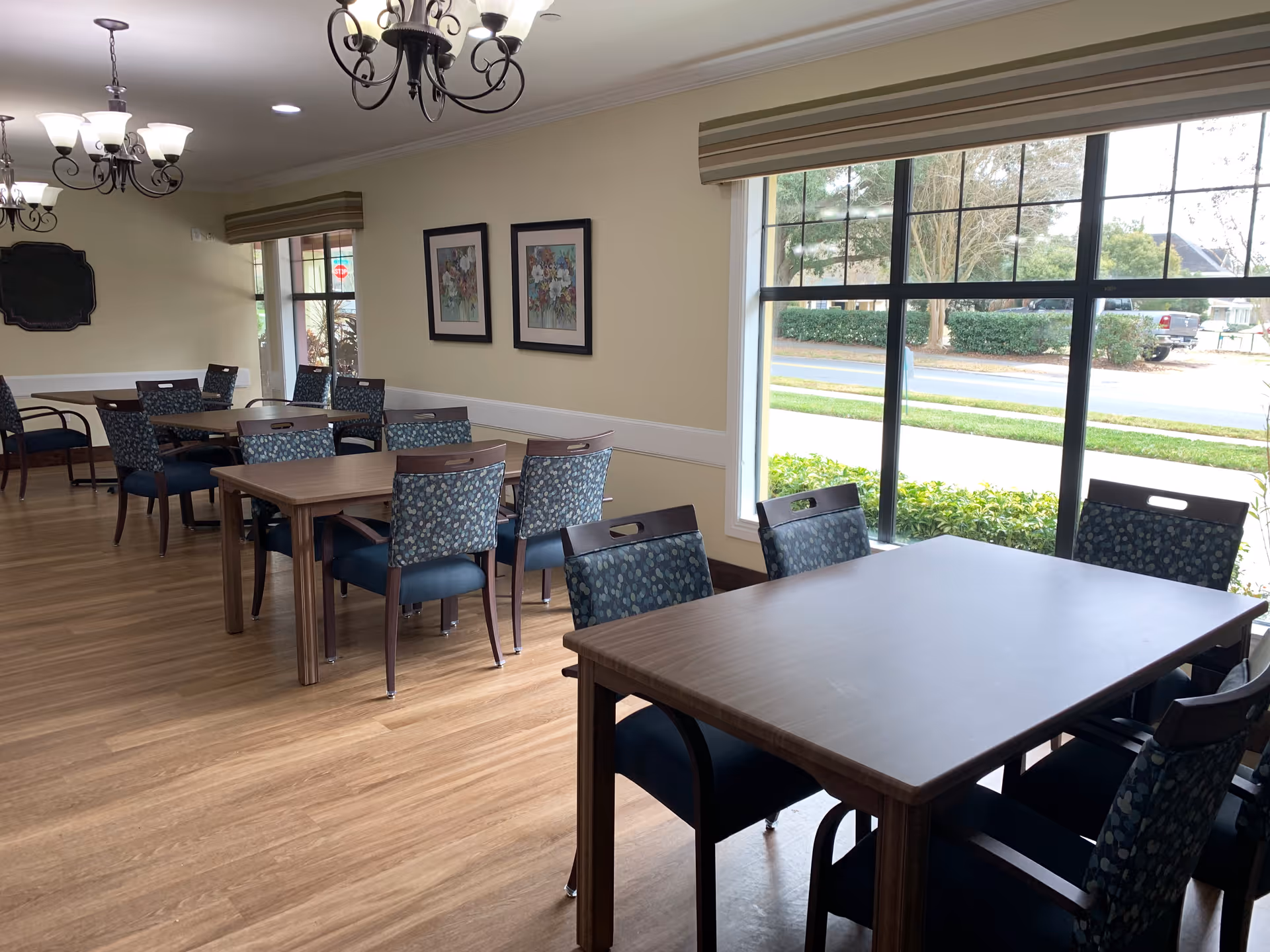 Bright dining room with multiple wooden tables and patterned chairs next to large windows overlooking landscaping.