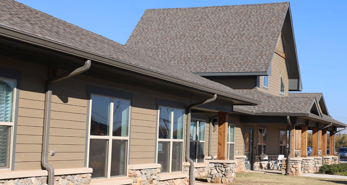 Exterior view of a senior assisted living facility building with beige siding, stone accents at the base, multiple windows, and a covered porch supported by wooden pillars under a clear blue sky.